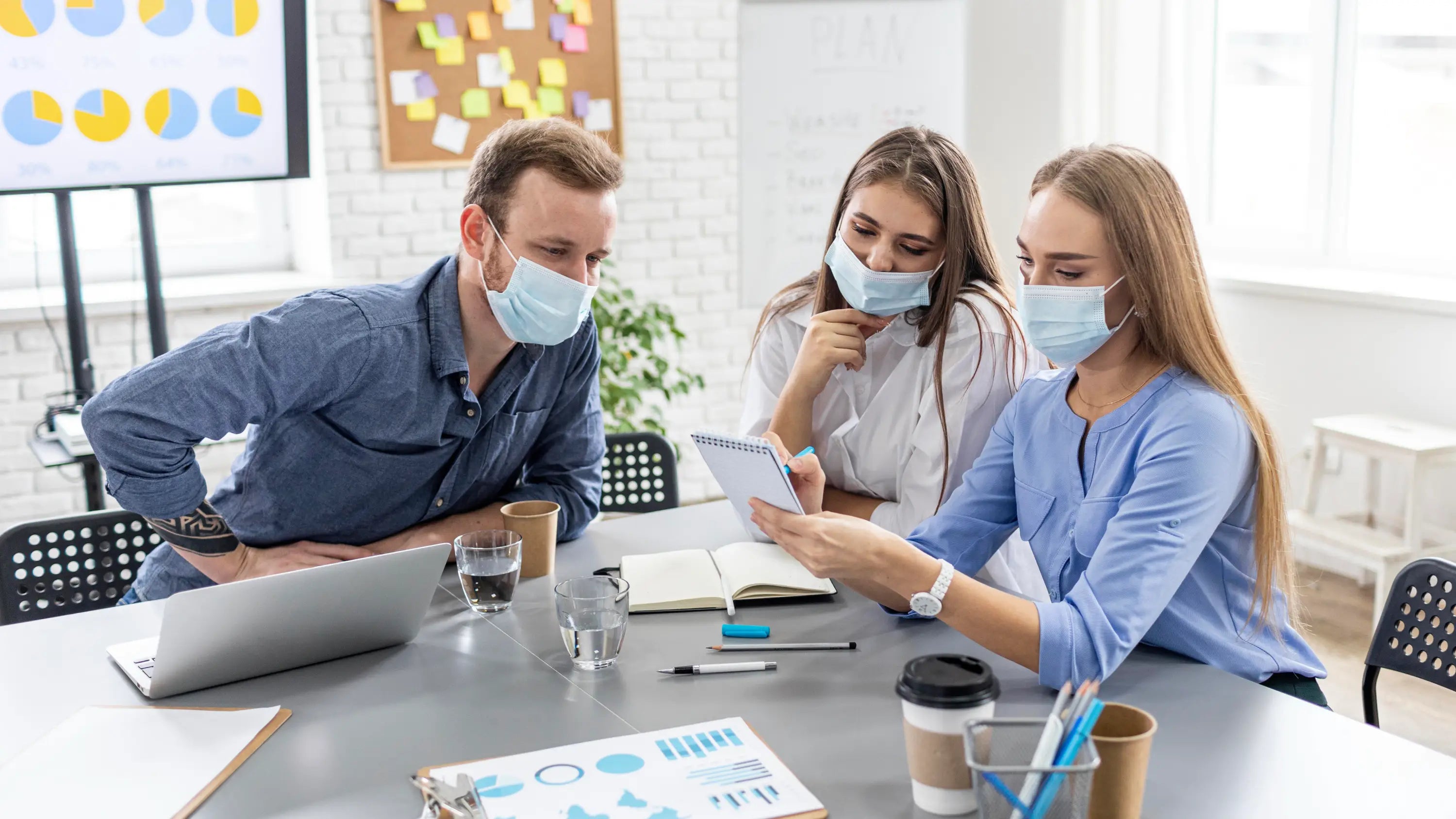 Three people in a modern office setting, wearing masks and working together.