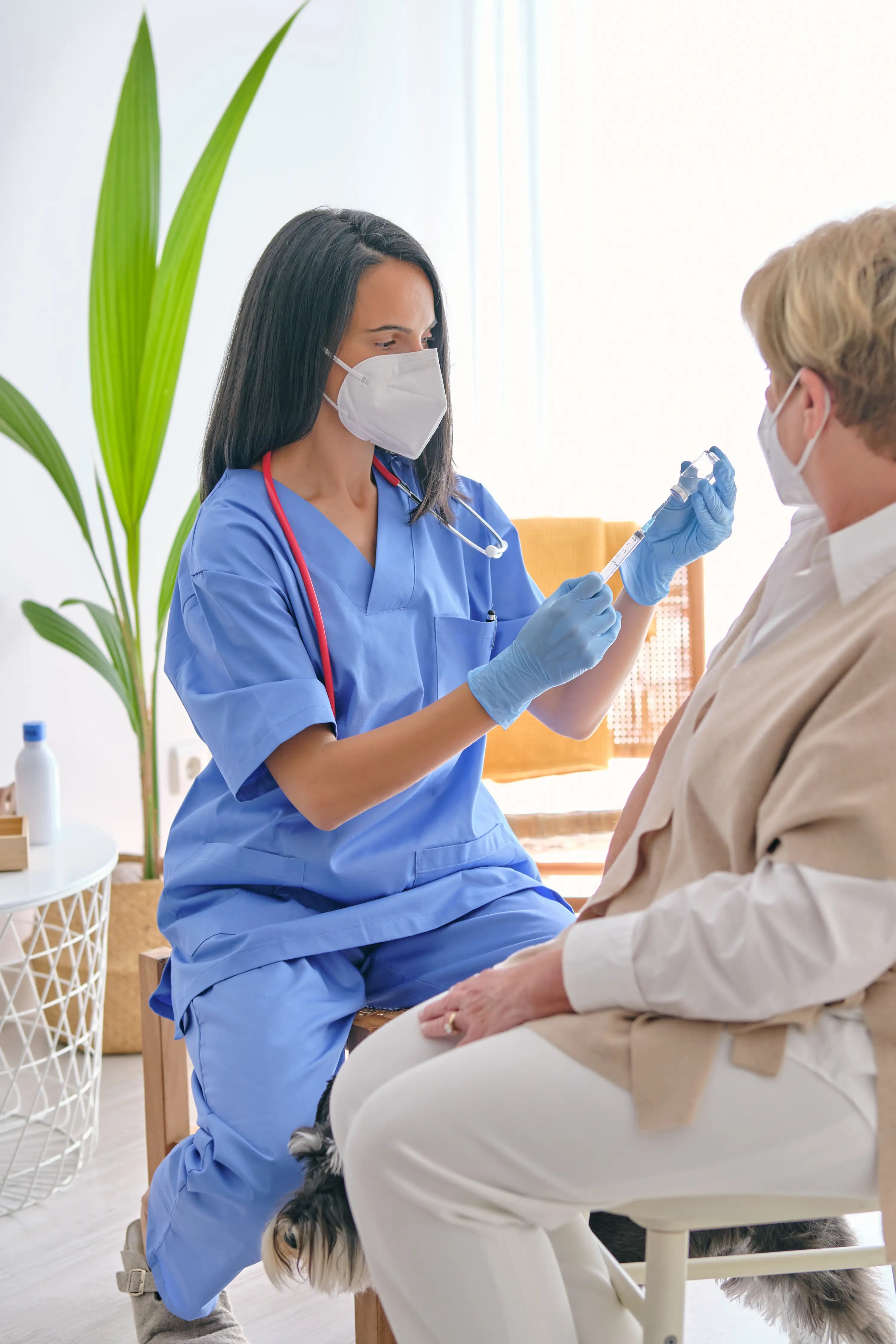 Nurse in blue scrubs and face mask examining a patient in a home setting.