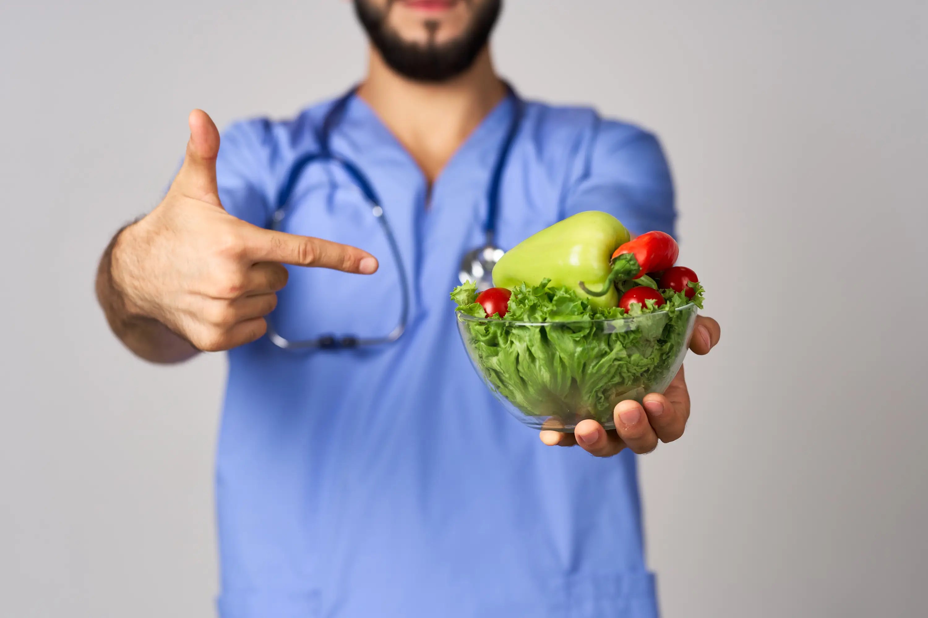 Person in blue scrubs pointing at a bowl of salad on a gray background