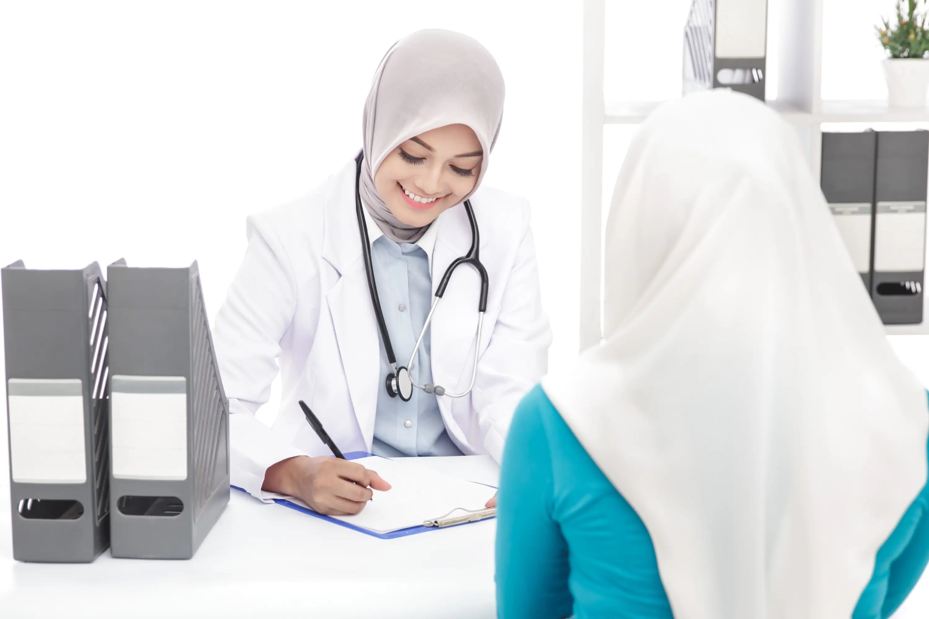 Doctor in white coat with stethoscope taking notes from a patient, both wearing hijabs.