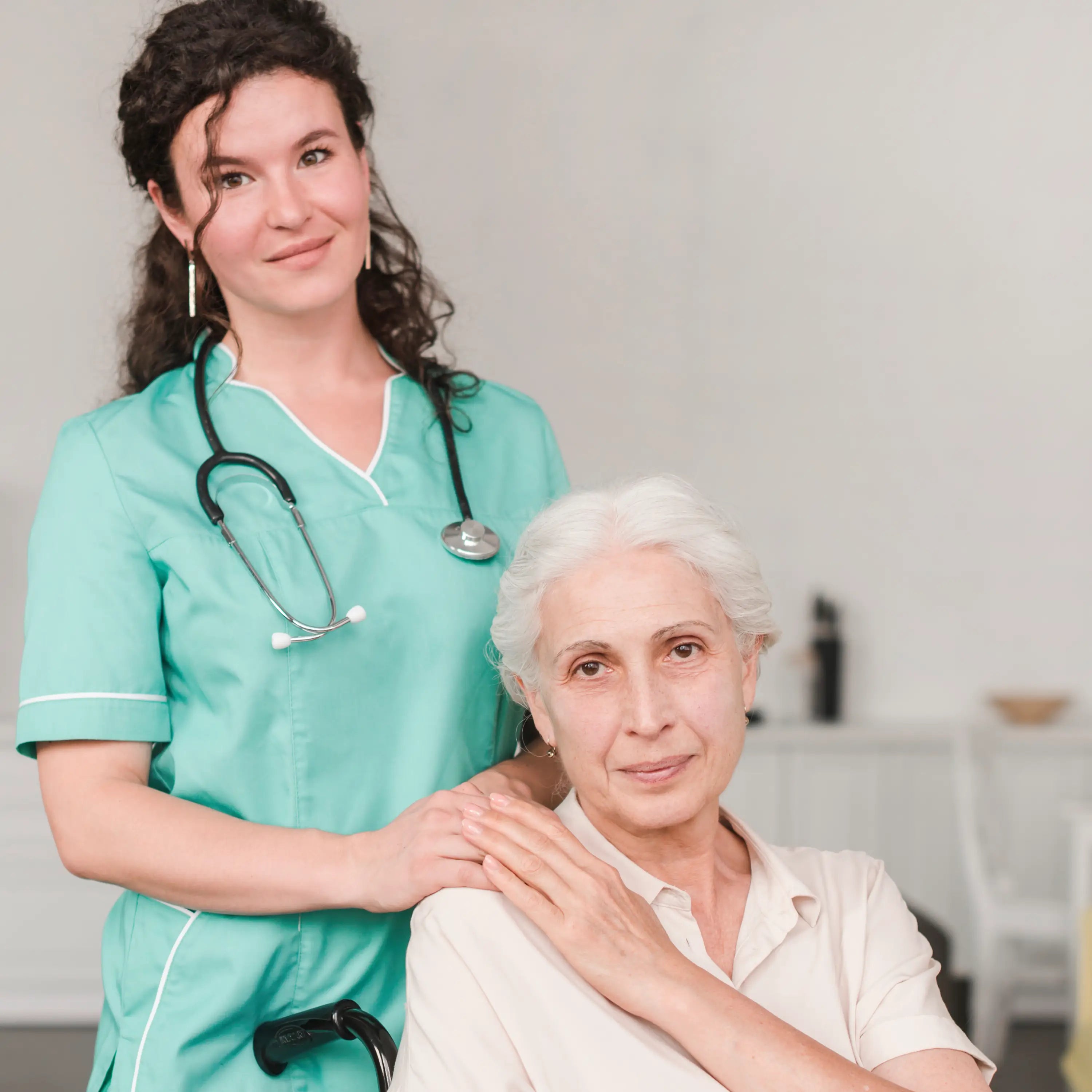 Nurse in teal scrubs with a stethoscope standing next to an elderly woman in a home setting.