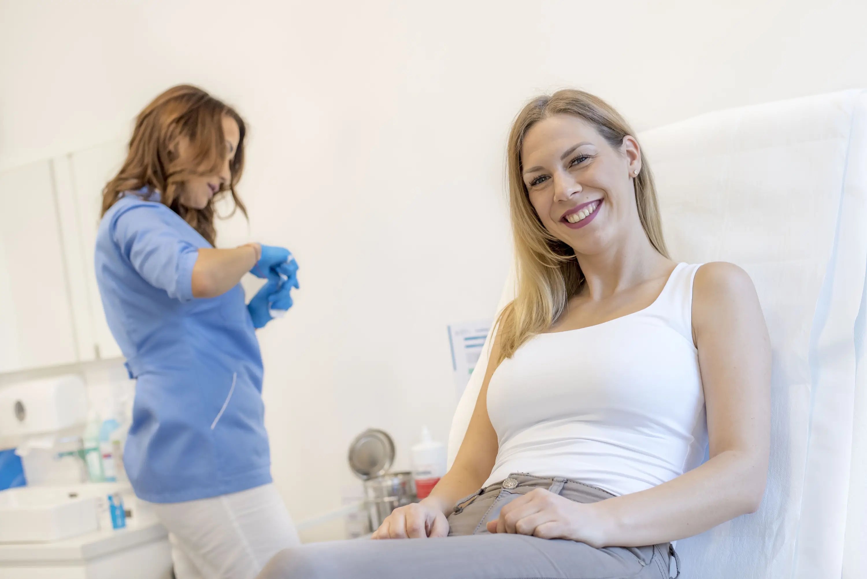 Woman sitting in a medical chair with a healthcare professional preparing for a procedure.
