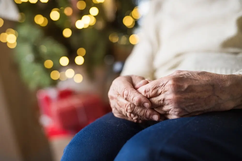 Close-up of an elderly woman's hands folded in her lap, sitting near a decorated Christmas tree