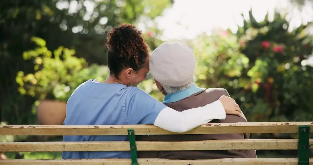 Caregiver sitting with a senior woman on a bench outdoors, sharing a warm hug