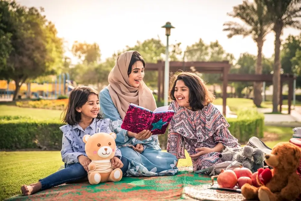 Mother and two daughters enjoying a picnic outdoors, reading a book together in a park