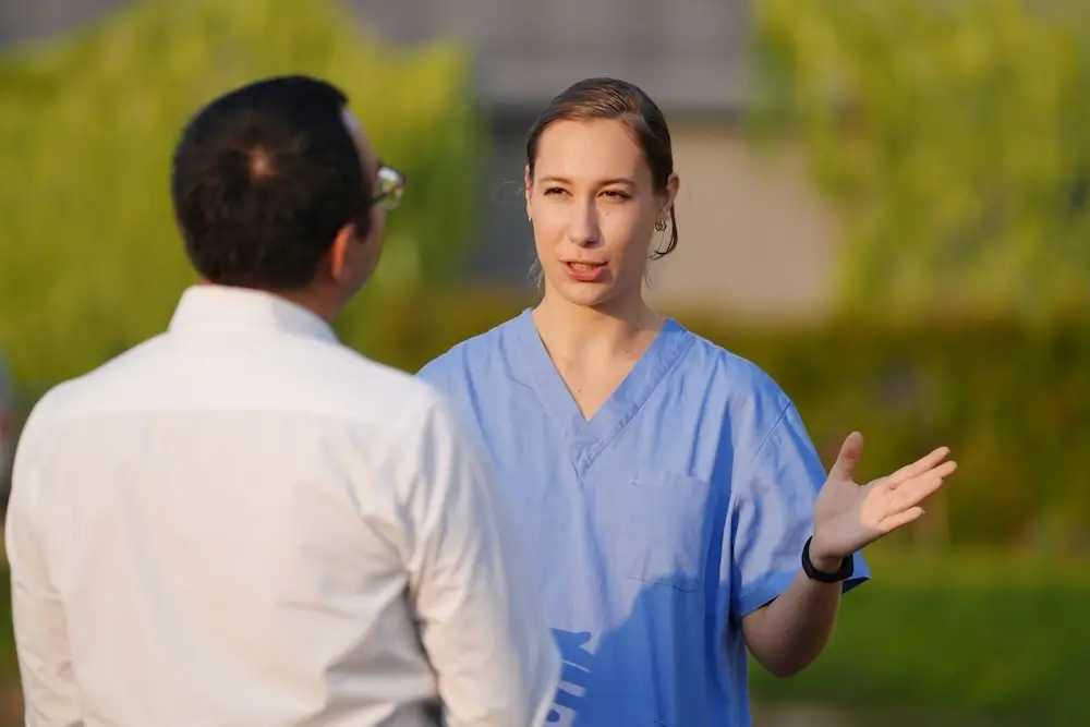 Female nurse in blue scrubs discussing care details with a colleague outdoors