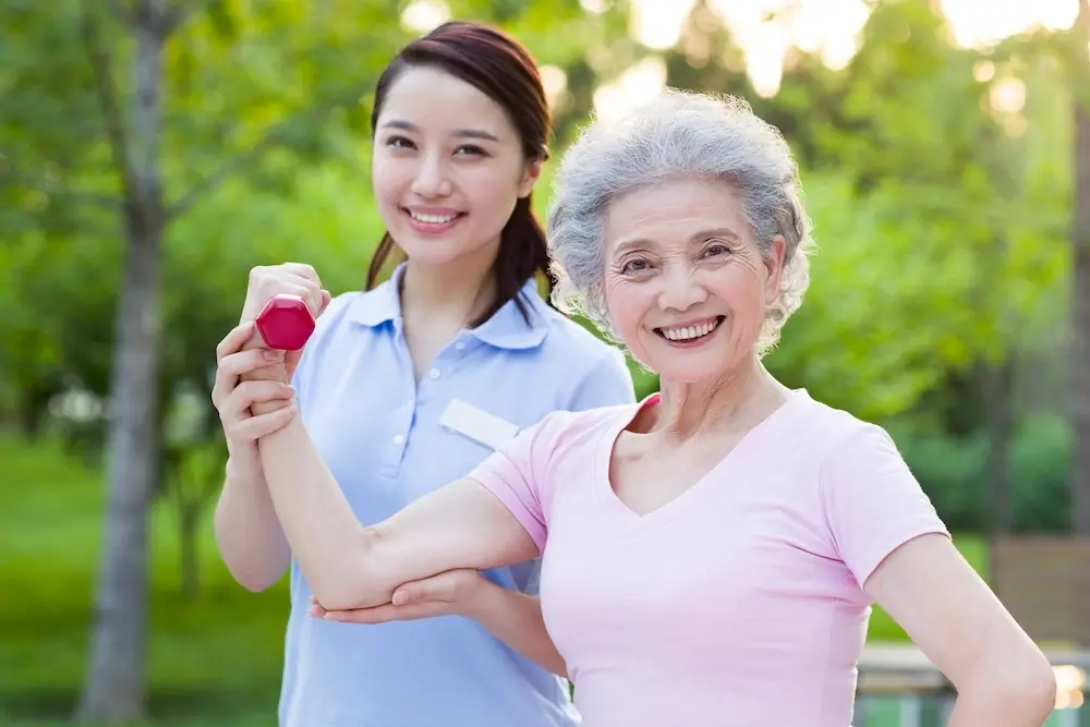 Smiling senior woman exercising with a dumbbell under the guidance of a caregiver, promoting active senior care in Qatar