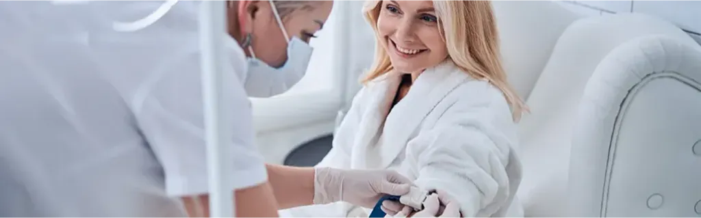 Smiling woman in a white robe receiving a blood draw from a medical professional wearing gloves and a mask in a clinical setting.